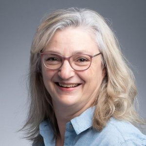 Photo of Sandi. A woman with long silver hair wearing glasses is smiling at the camera.